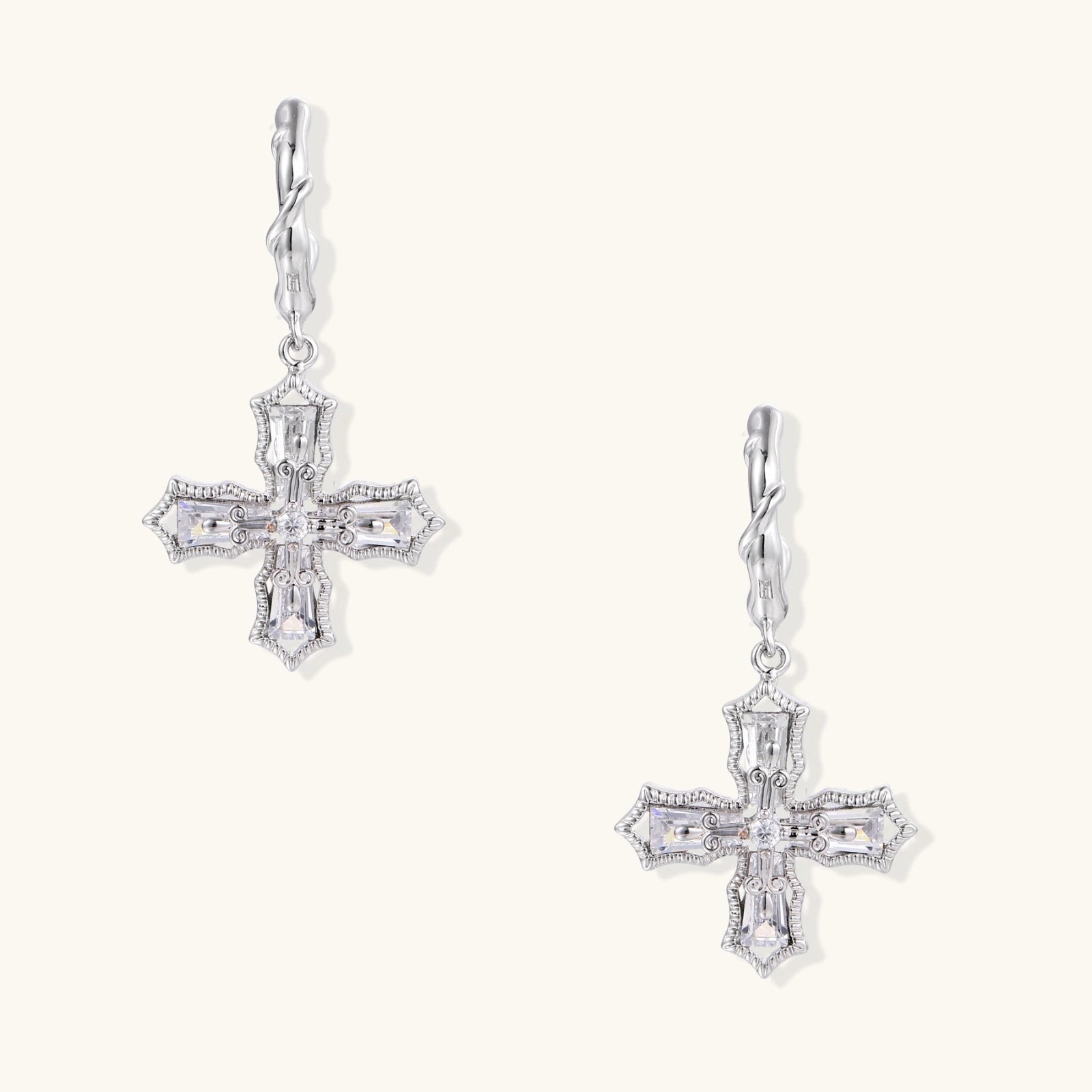 Ornate white gold-plated cross earrings with baguette-cut zirconia stones and twisted leverback hooks on beige background