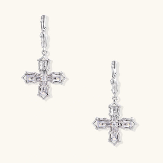Ornate white gold-plated cross earrings with baguette-cut zirconia stones and twisted leverback hooks on beige background